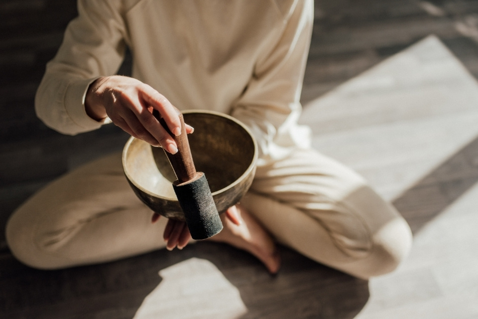 faceless person holding out singing bowl in front of them while sitting crossed legs on wooden floor afternoon sun