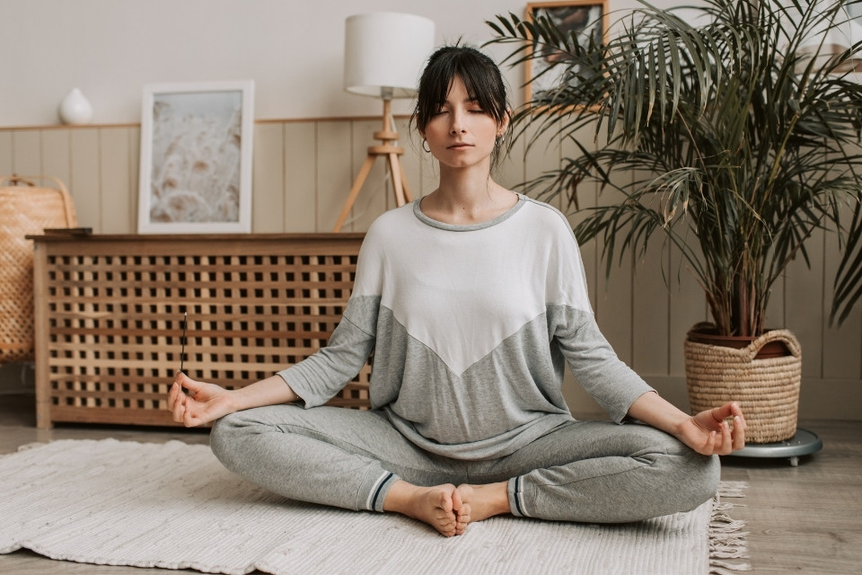 woman crossed legs while meditating on wooden floor with gray rug indoor with plant, furniture and household decors